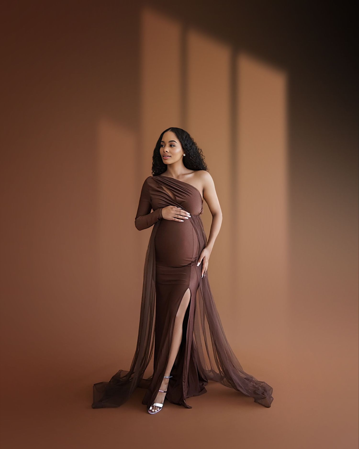 Elegant maternity photo of a woman in a flowing brown gown posing against a neutral backdrop with dramatic window lighting.