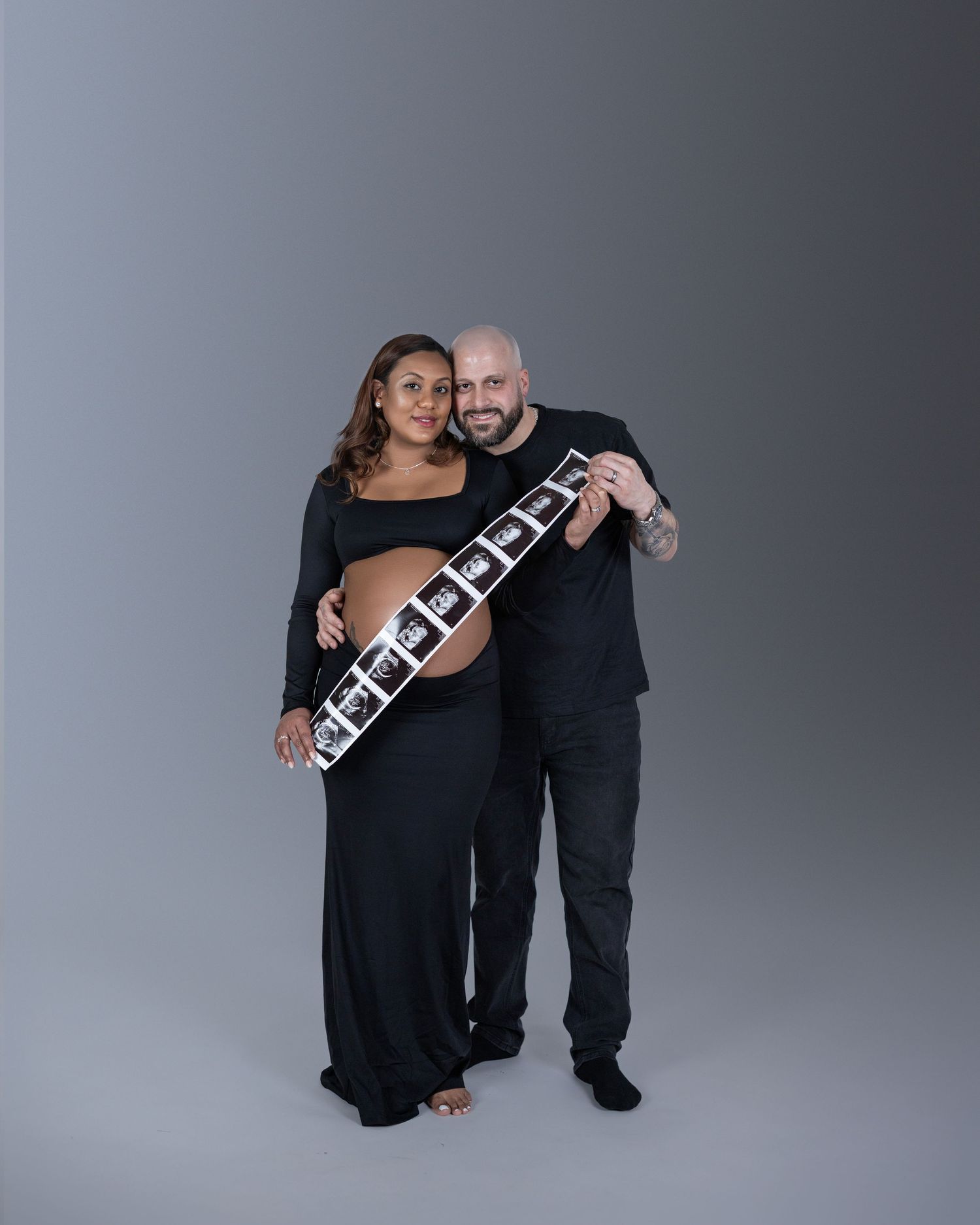 Two people in black outfits pose together against a gray background with a decorative sash or banner.