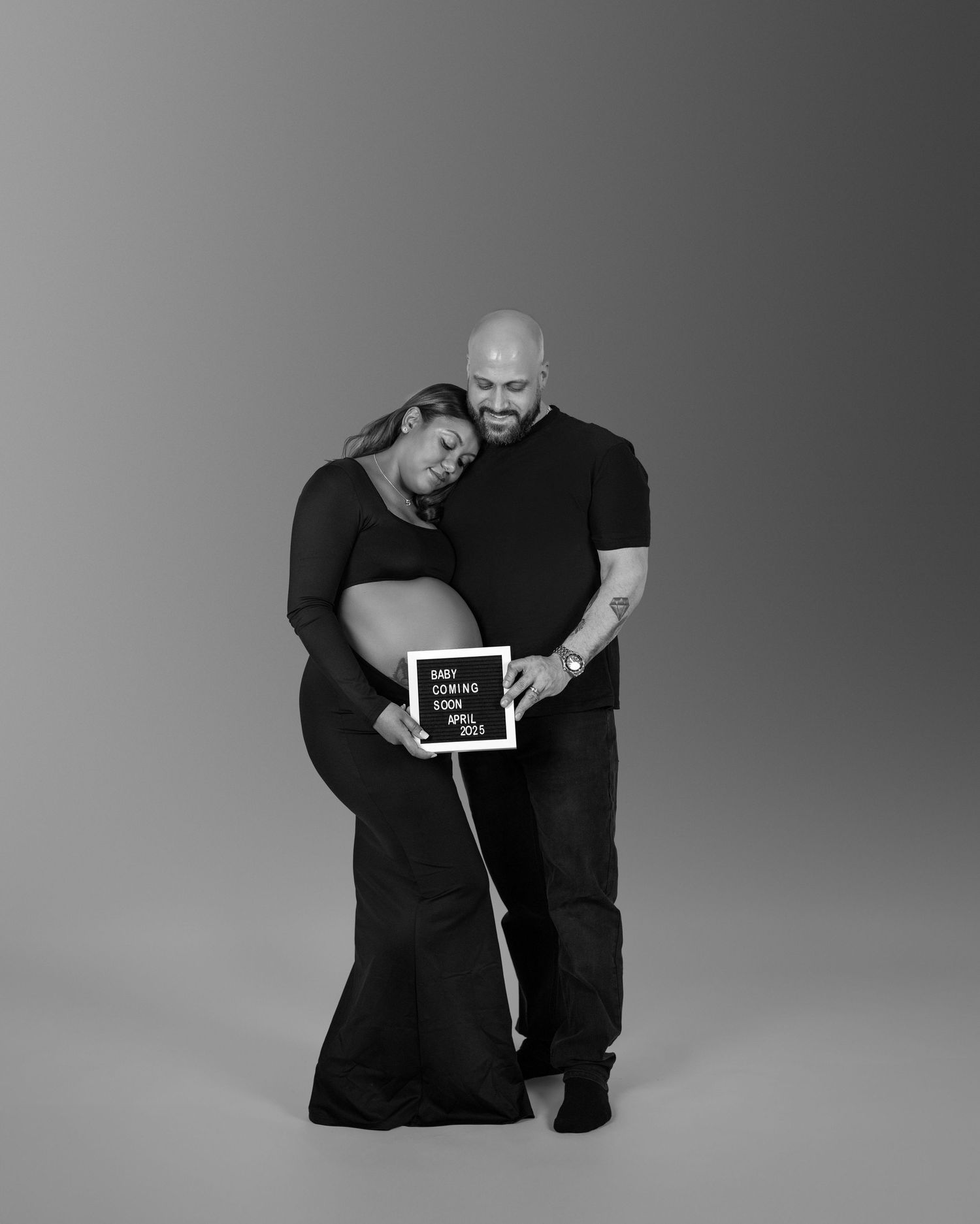 Black and white studio photo showing a couple in dark clothing holding an ultrasound image during a pregnancy announcement.