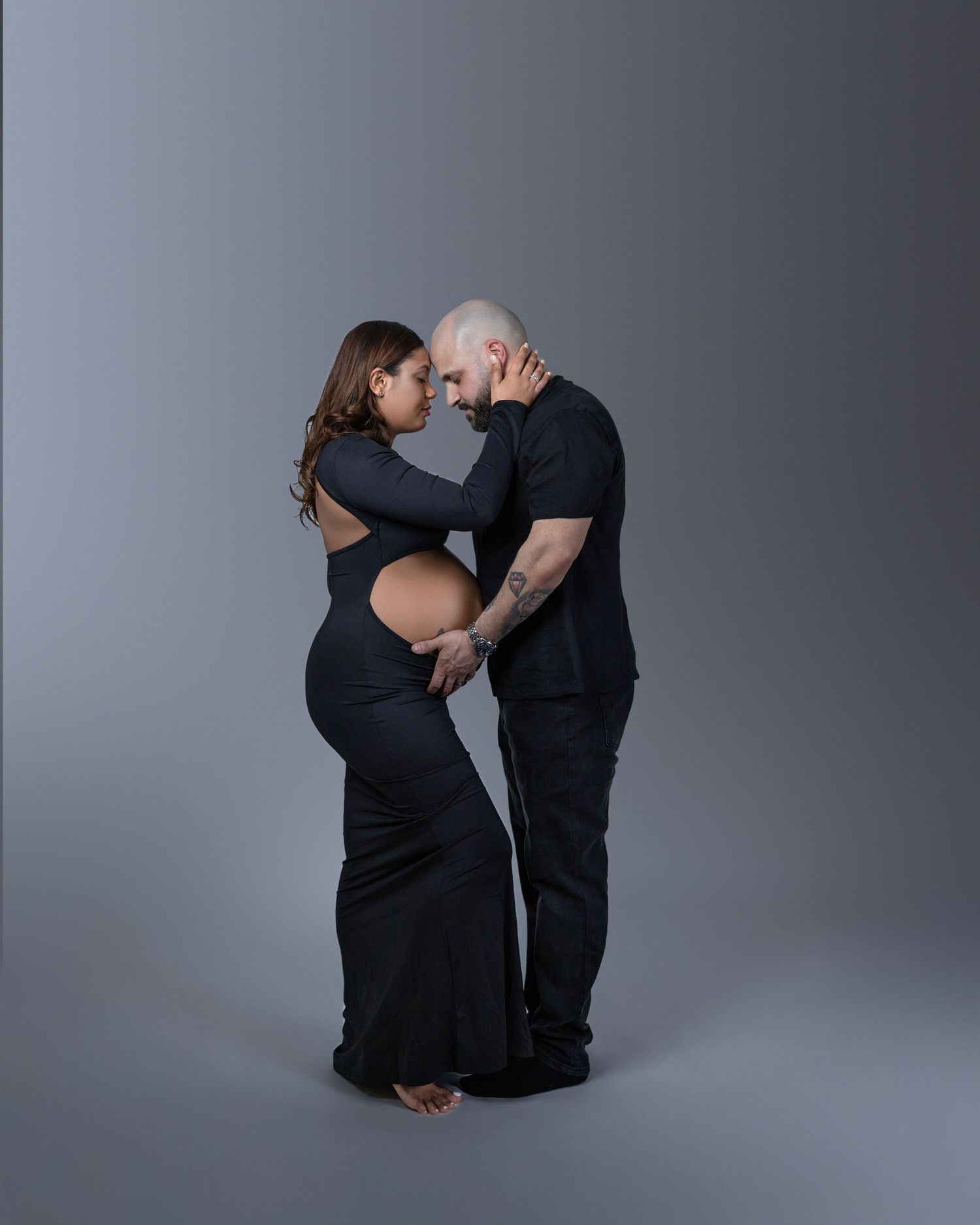 Intimate maternity portrait of a couple wearing black embracing against a gray studio backdrop.