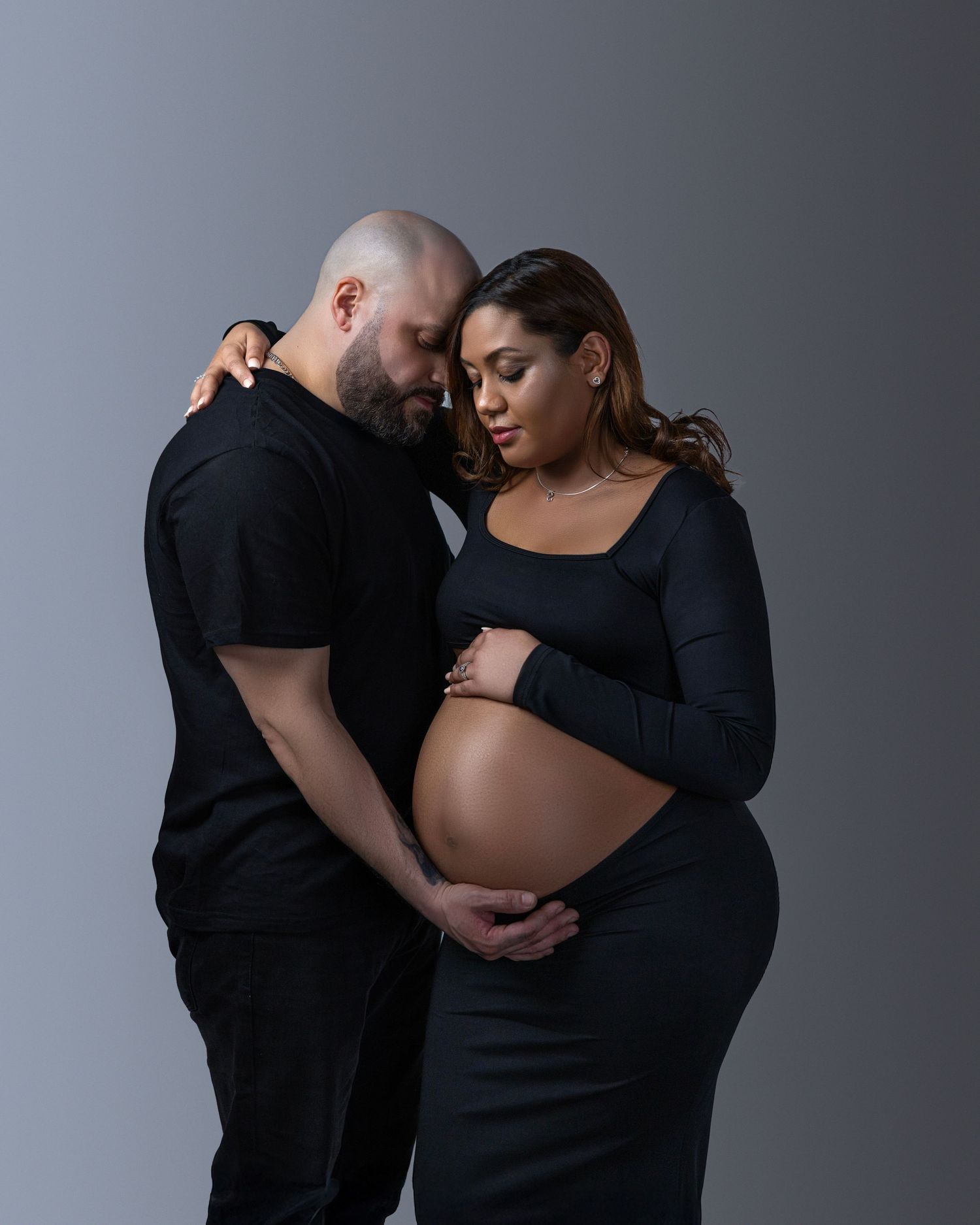 Intimate maternity photo of expectant couple in black outfits embracing against gray background.