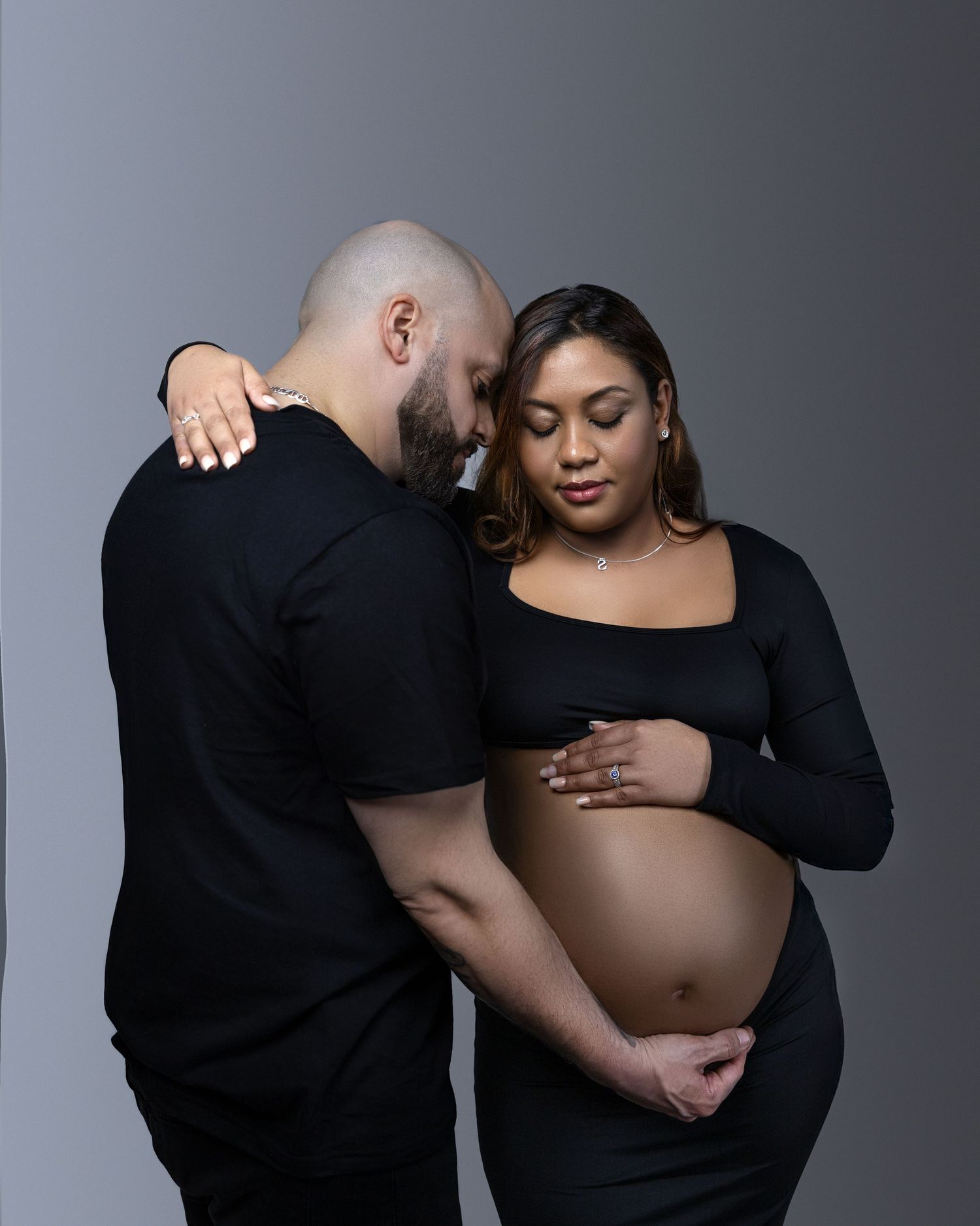 A couple shares an intimate moment during a maternity photoshoot against a gray studio backdrop.