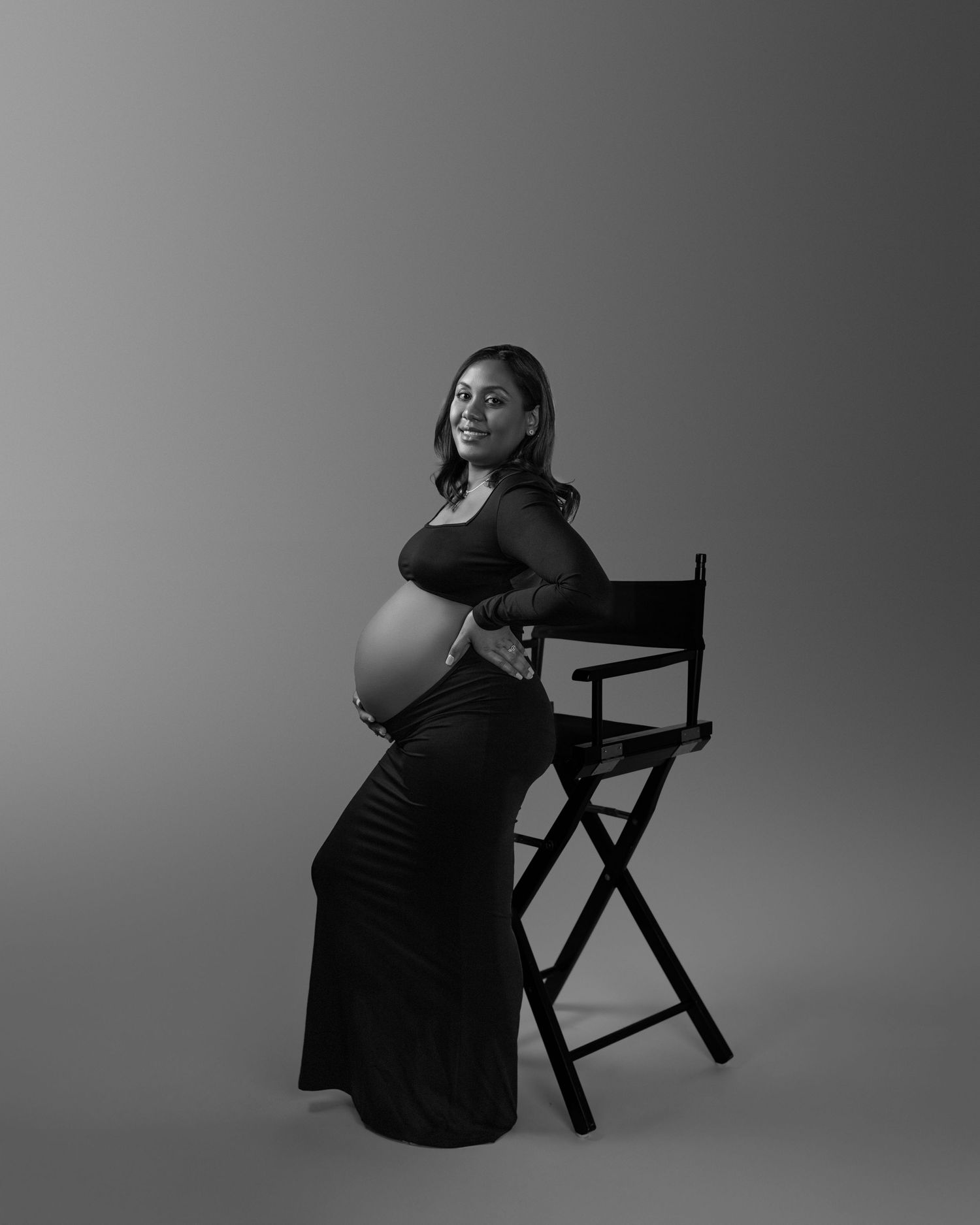 A pregnant woman in black dress poses elegantly against a dark chair in an artistic black and white studio portrait.