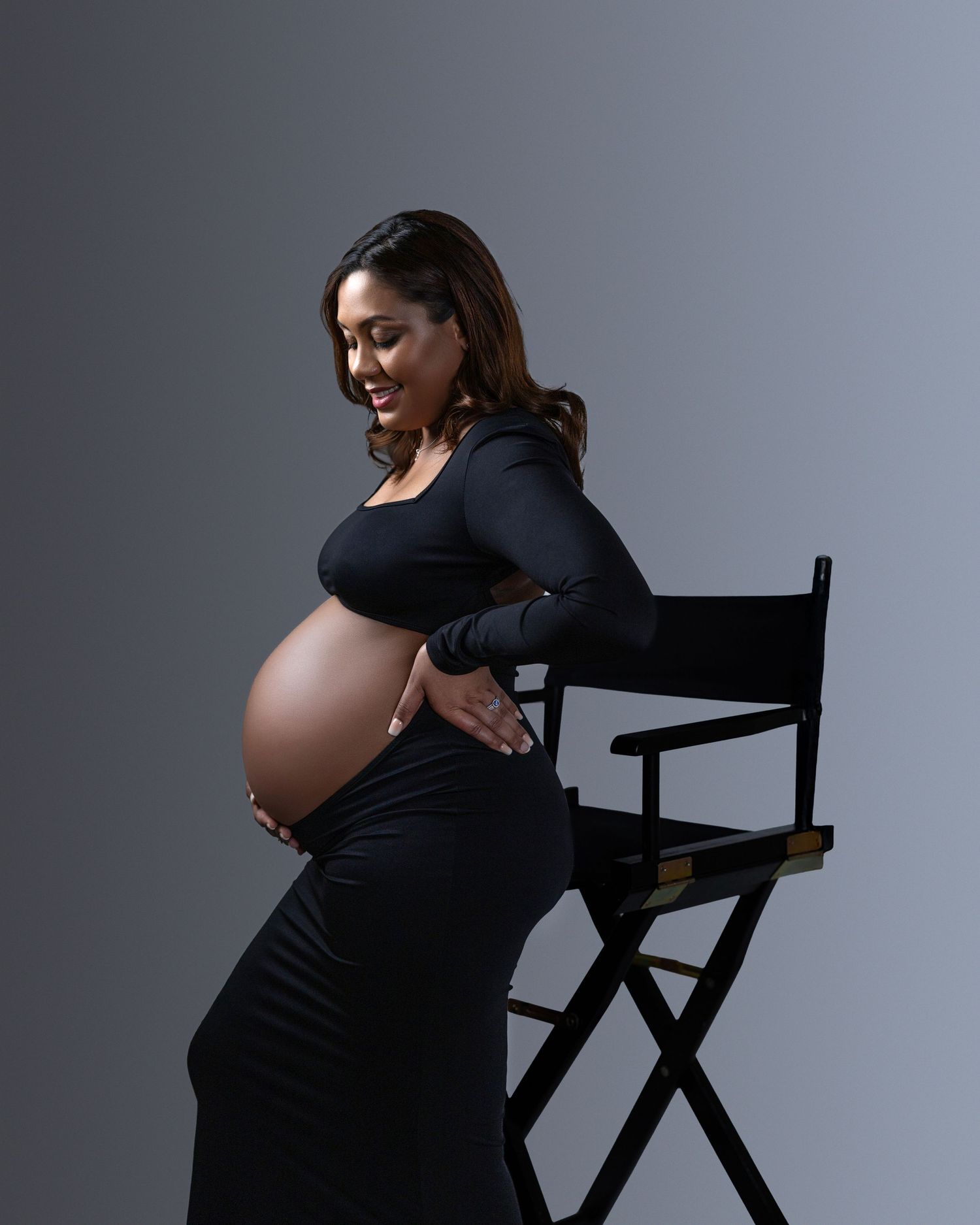 A pregnant person in a black dress poses elegantly next to a director's chair against a gray studio backdrop.