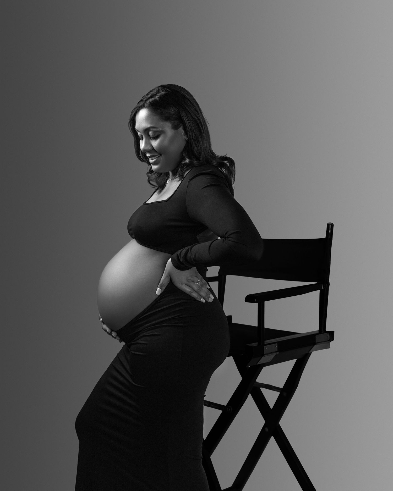 A black and white artistic maternity portrait showing an expectant mother in a fitted black dress seated against a chair.