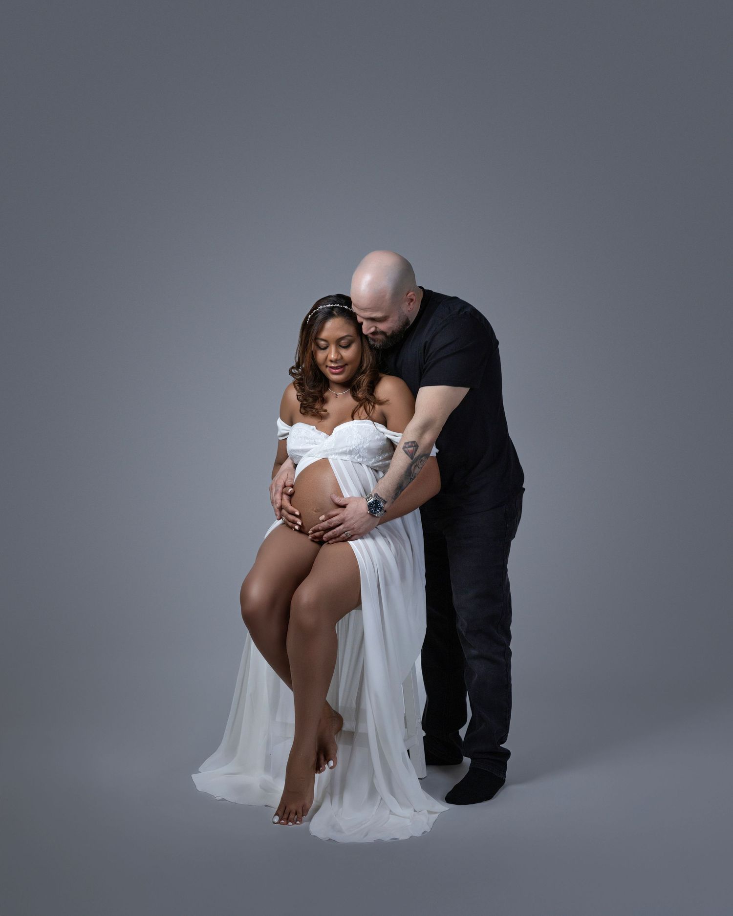 Intimate maternity portrait featuring an expecting couple in white flowing fabric against a grey studio backdrop.