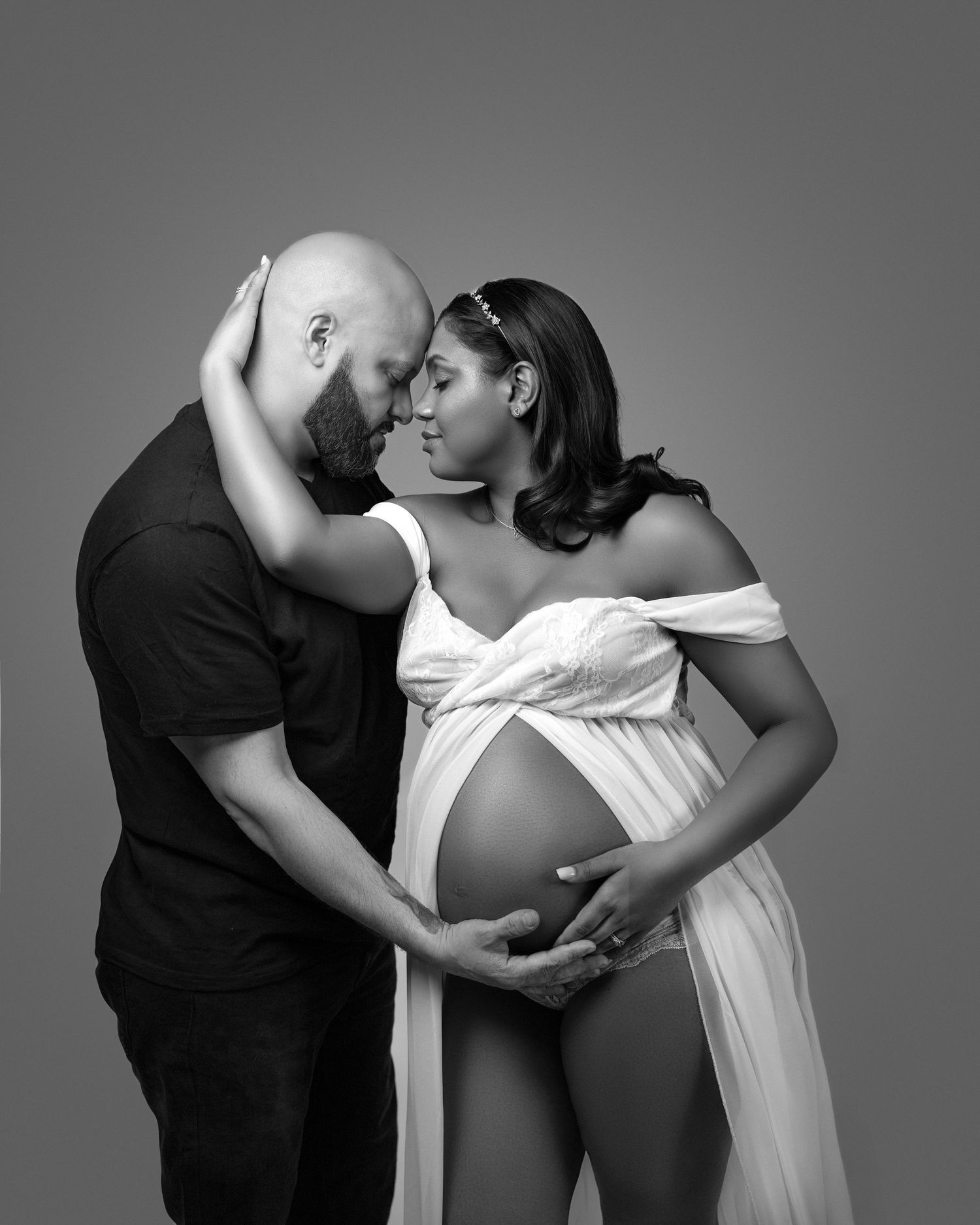 Black and white maternity photo showing intimate moment between couple in flowing white dress against grey backdrop.