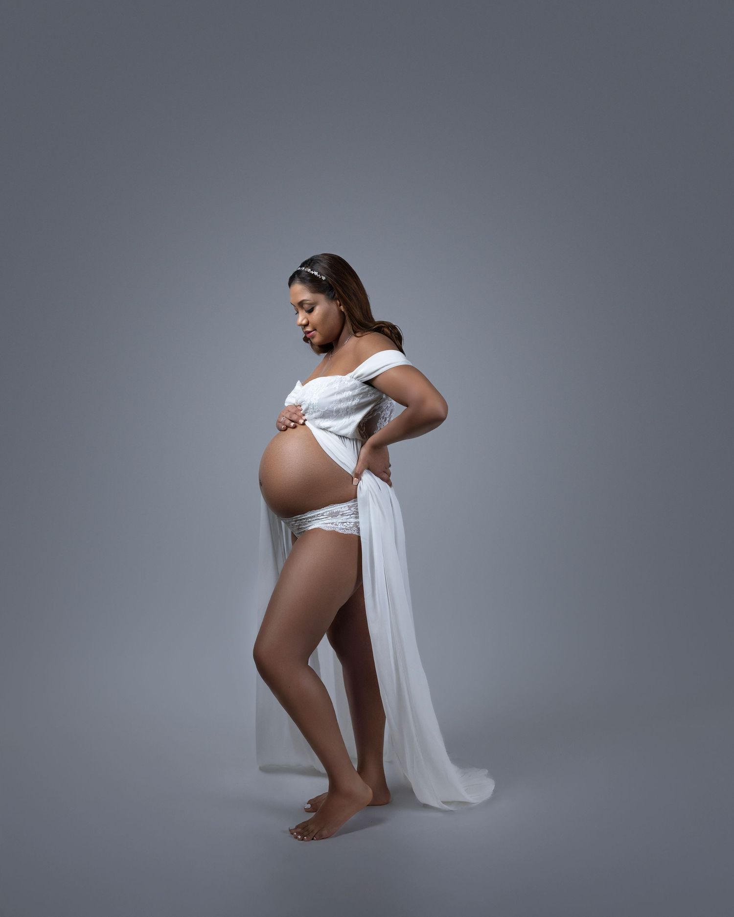 Ethereal maternity portrait with flowing white fabric draping around expectant mother against grey background.