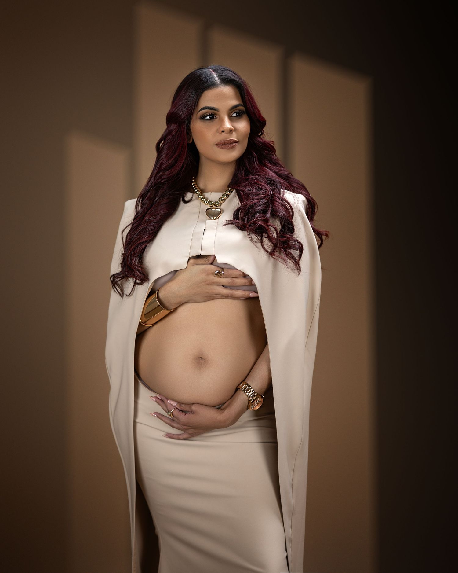 Maternity photo in soft lighting shows expectant mother in white dress and cape against dark wall with dramatic shadows.