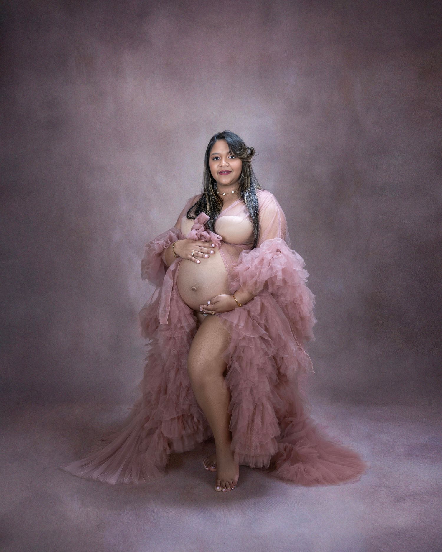A pregnant woman poses in a flowing dusty rose tulle gown against a moody studio backdrop.