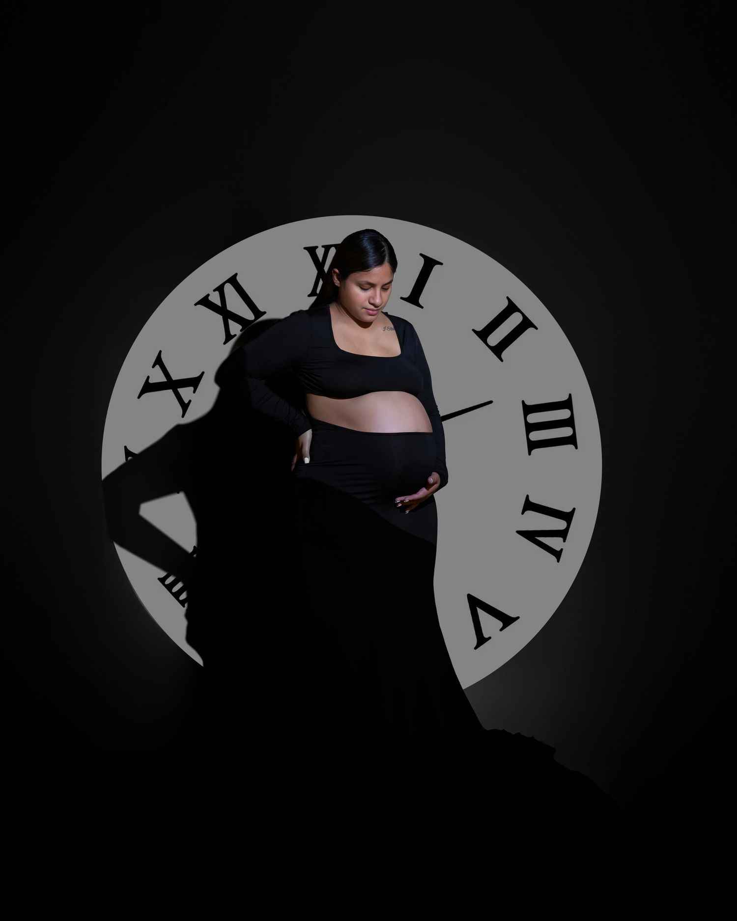 Silhouetted figure in dark clothing poses artistically against a large illuminated clock face in a dramatic maternity photo.