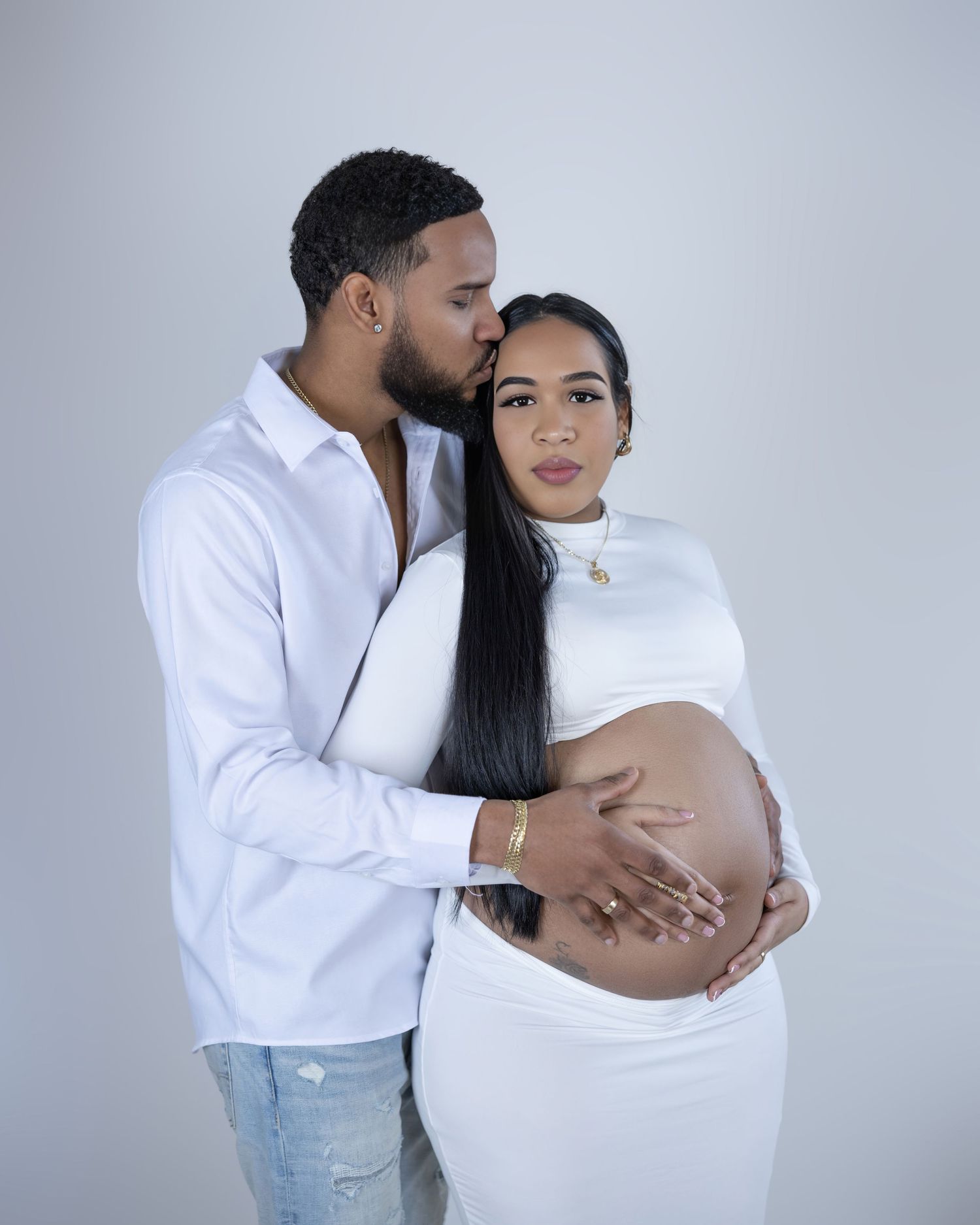 A couple in white outfits embraces during a maternity photoshoot against a gray background.