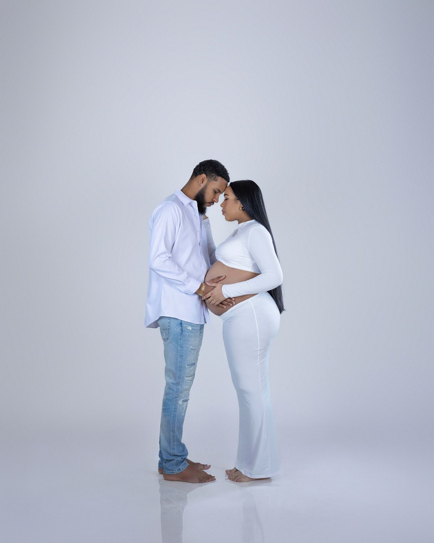 Expectant couple in matching white outfits share an intimate moment against a gray studio backdrop.