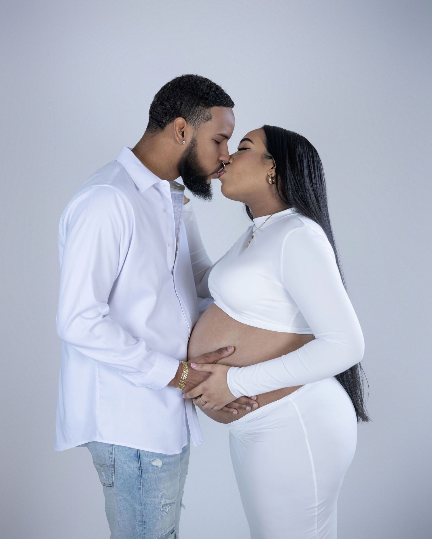 A couple in matching white outfits share an intimate moment during a maternity photoshoot against a gray backdrop.