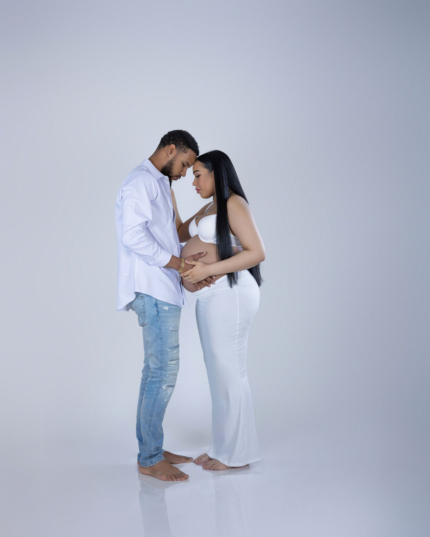 A couple in white clothing share an intimate moment standing barefoot against a light gray background.