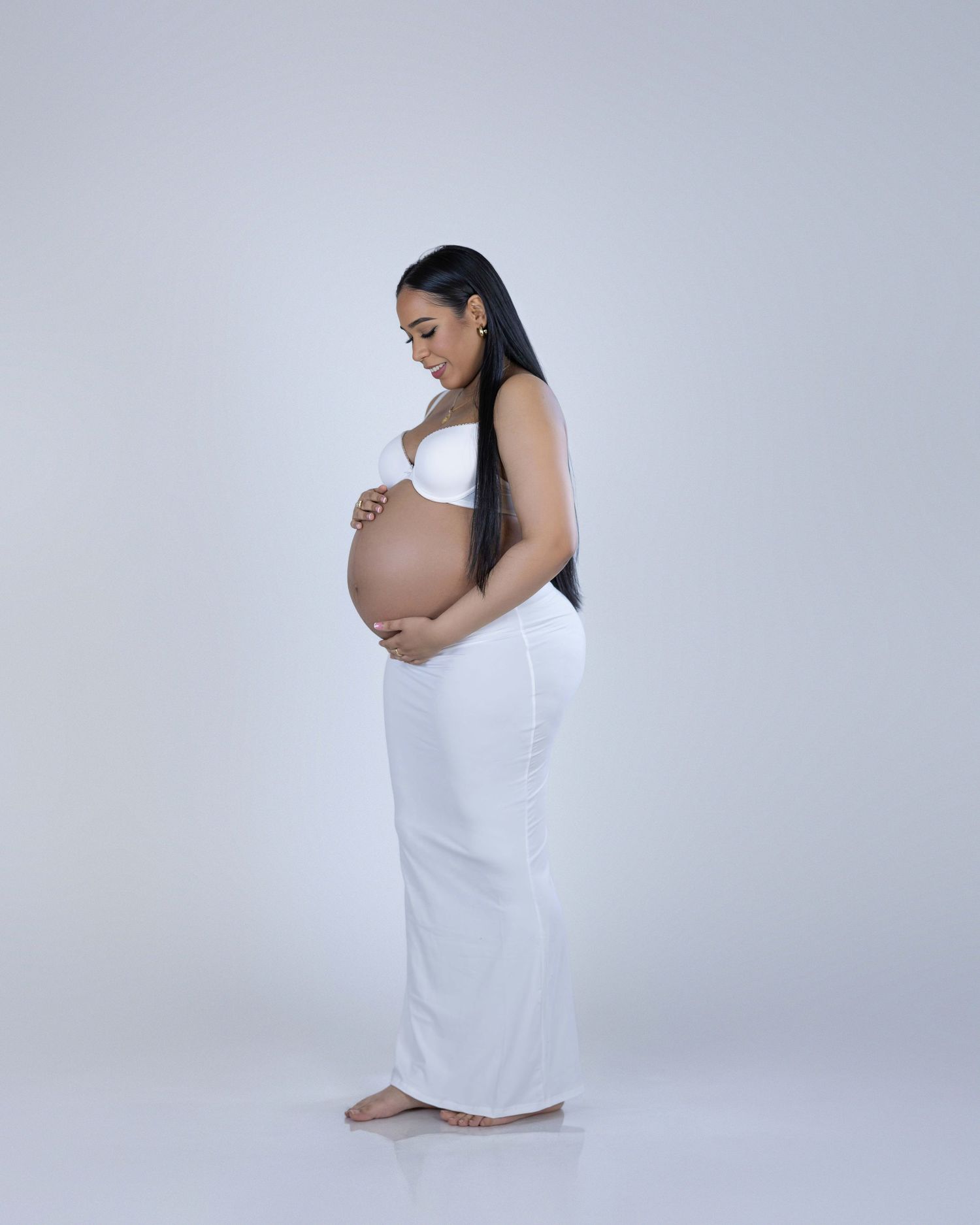 A pregnant woman in a flowing white dress poses against a gray backdrop while cradling her belly.