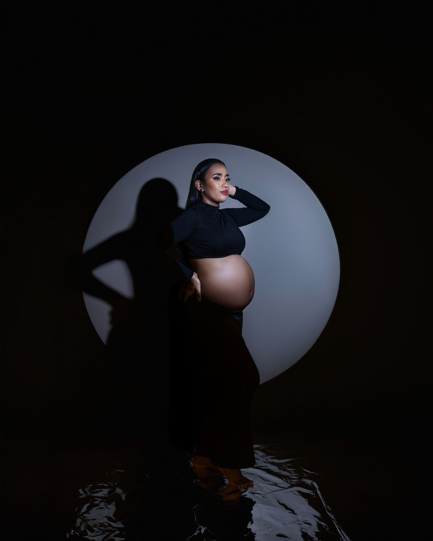 Artistic silhouette maternity photo with a pregnant figure against a circular light backdrop in dramatic lighting.