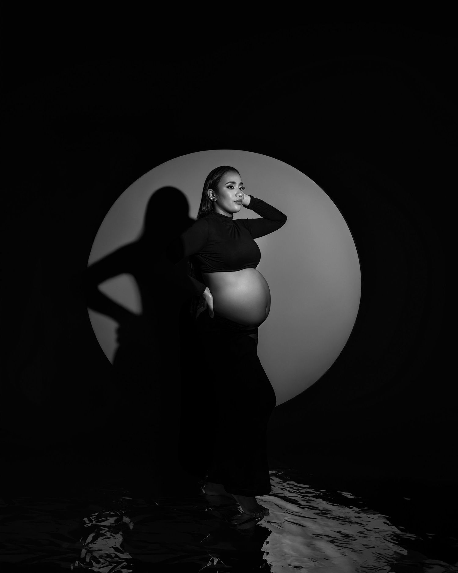 Artistic black and white maternity photo with dramatic circular lighting and shadow against dark background.