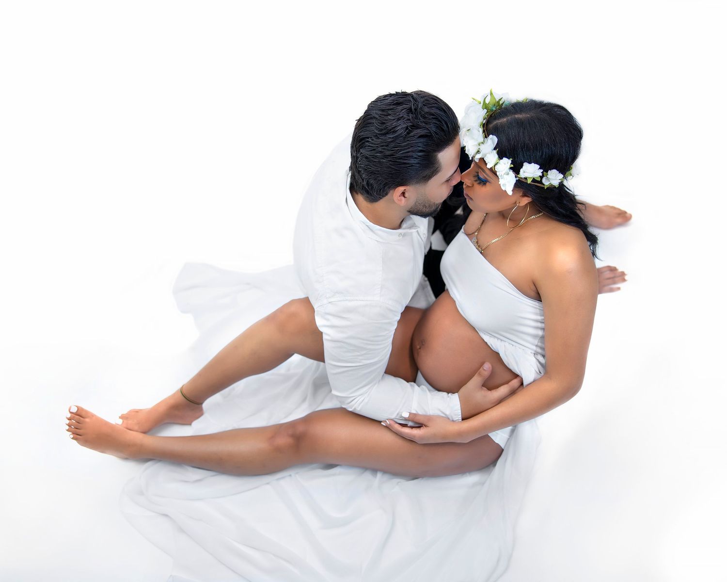 Intimate couple in white clothing embrace tenderly in soft wedding-style portrait against white background.