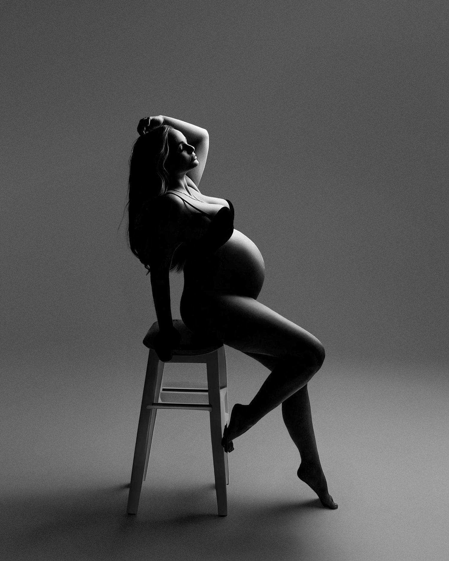 Black and white artistic profile silhouette of pregnant figure posing elegantly on a stool, highlighting maternity curves.