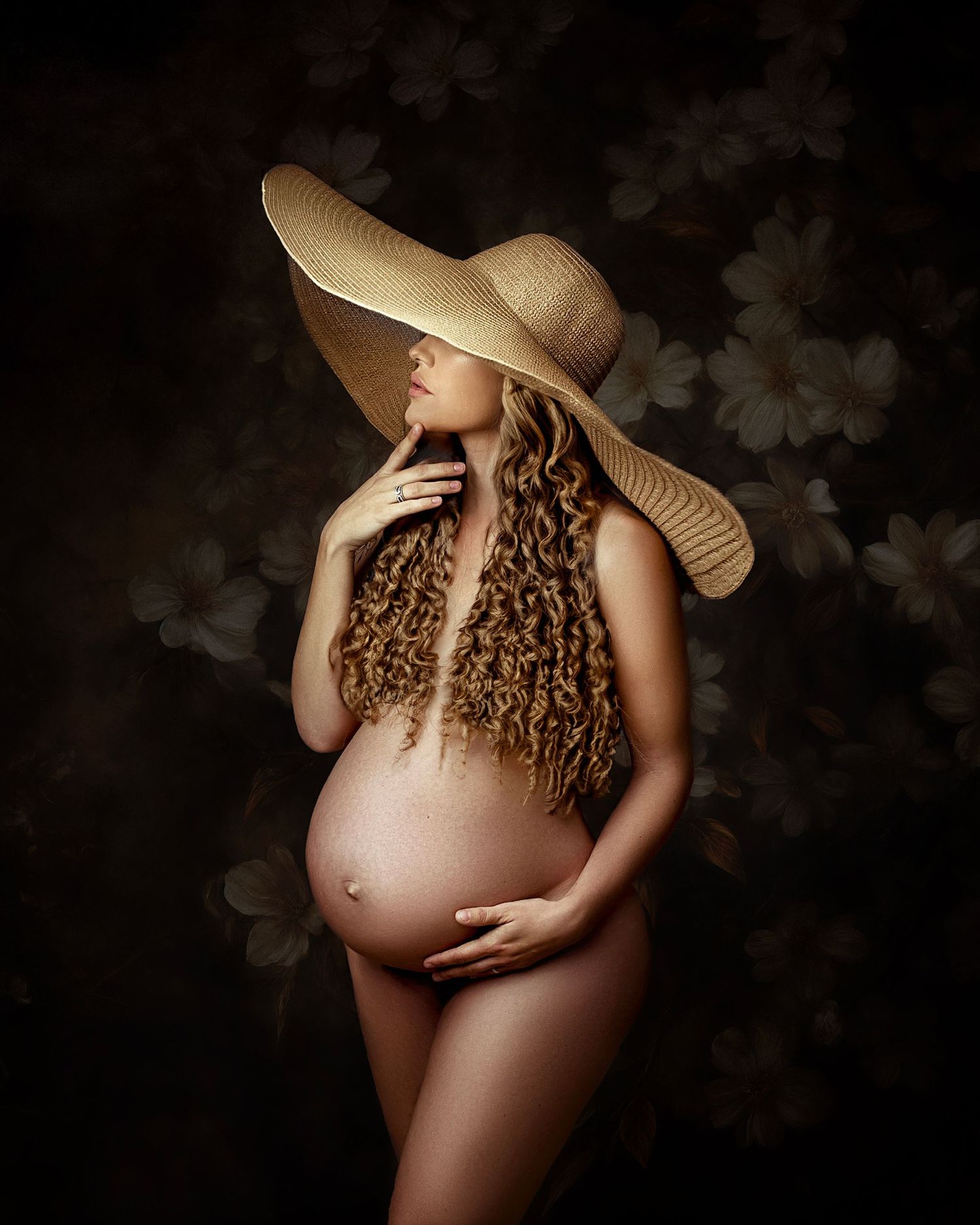 Artistic maternity photo featuring a nude figure in soft lighting wearing a large floppy sun hat with flowing curly hair.