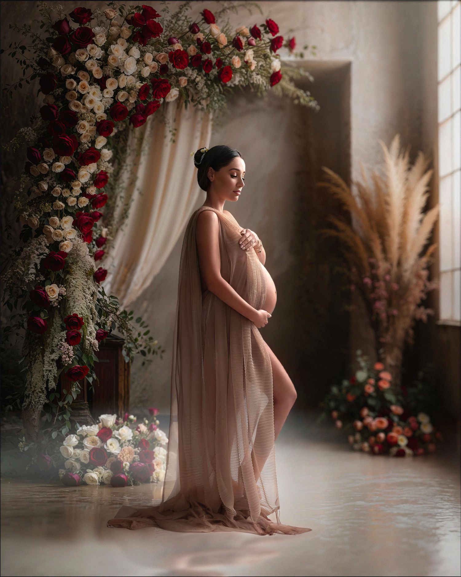 Romantic maternity photoshoot featuring floral arch with red and white roses, pampas grass, and flowing blush gown.