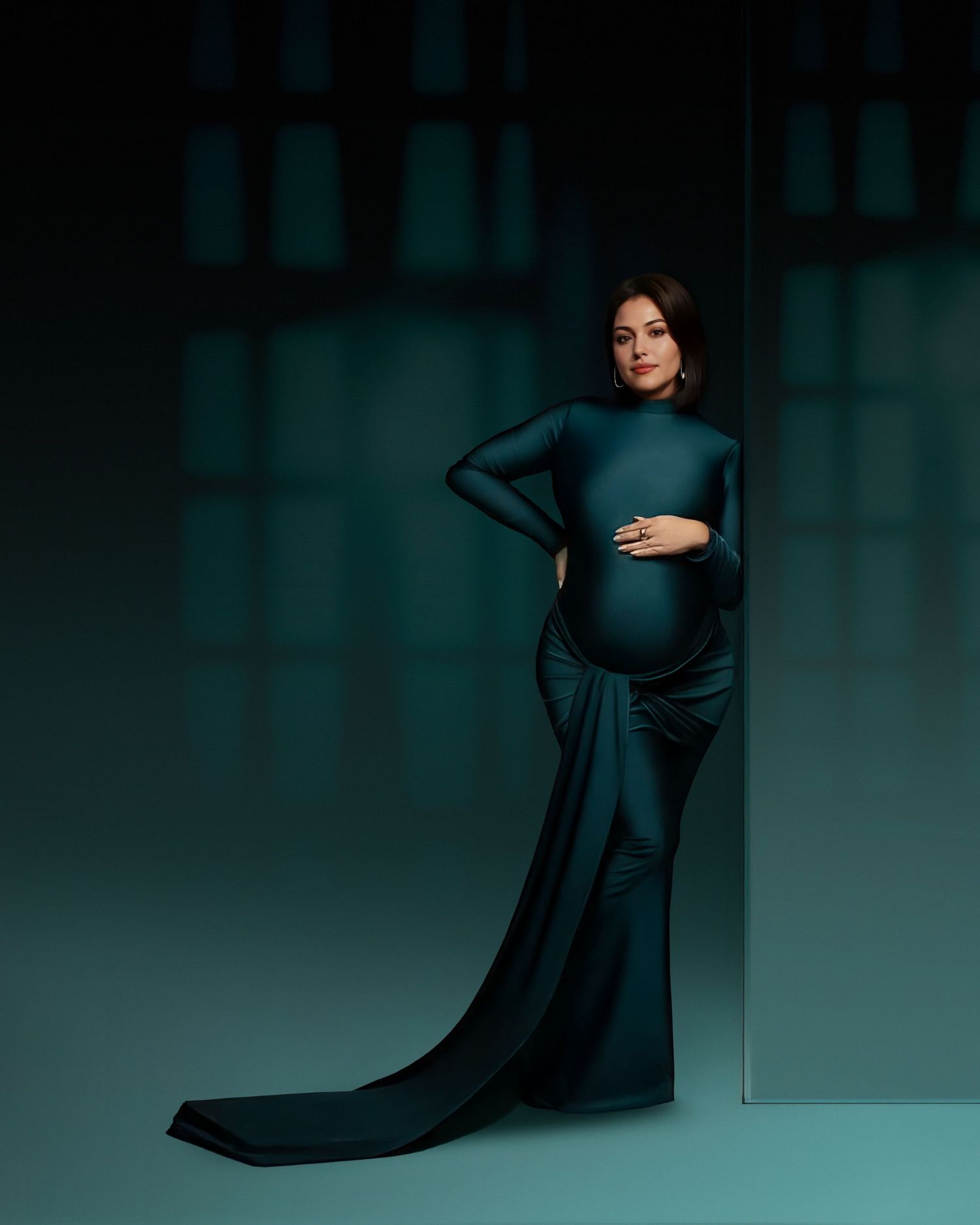 Artistic maternity photo with a woman in a flowing teal gown against a dark teal background with dramatic lighting.