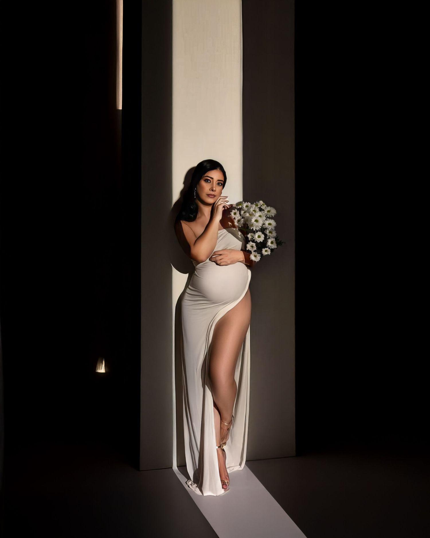An expecting mother poses in a fitted white dress with a thigh-high slit against a dramatically lit backdrop with floral decor.
