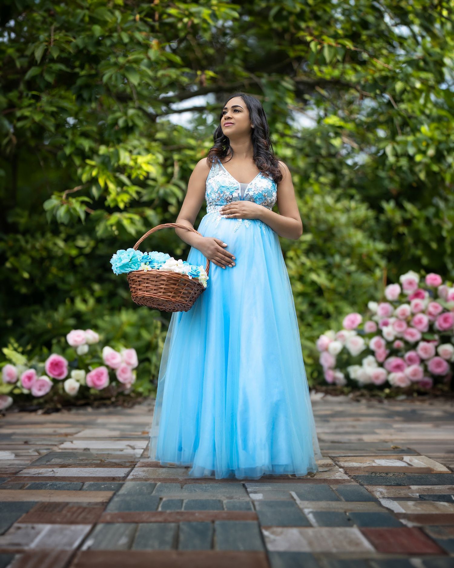 A person in a flowing light blue gown poses in a garden setting with pink roses and greenery on a brick pathway.