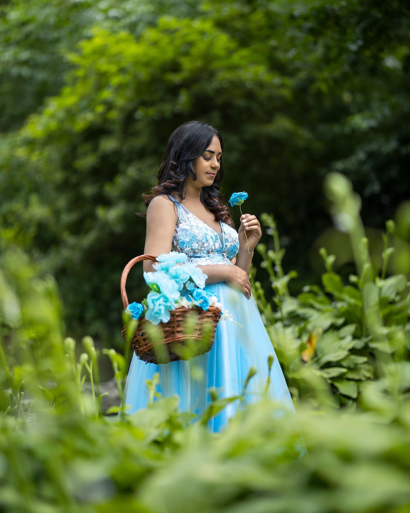 Figure in a light blue dress sits elegantly in a garden setting holding a basket filled with flowers.