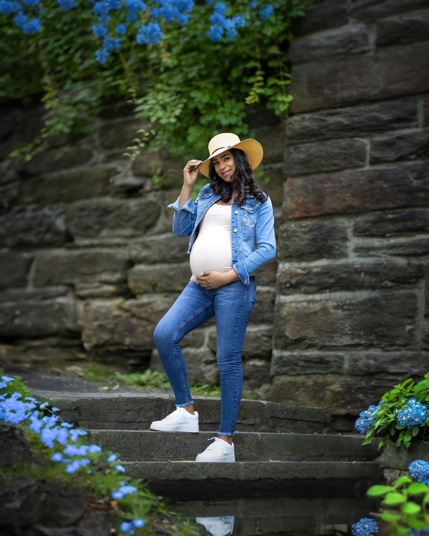 A pregnant woman in denim and blue jacket poses on stone steps surrounded by blue hydrangeas and greenery.