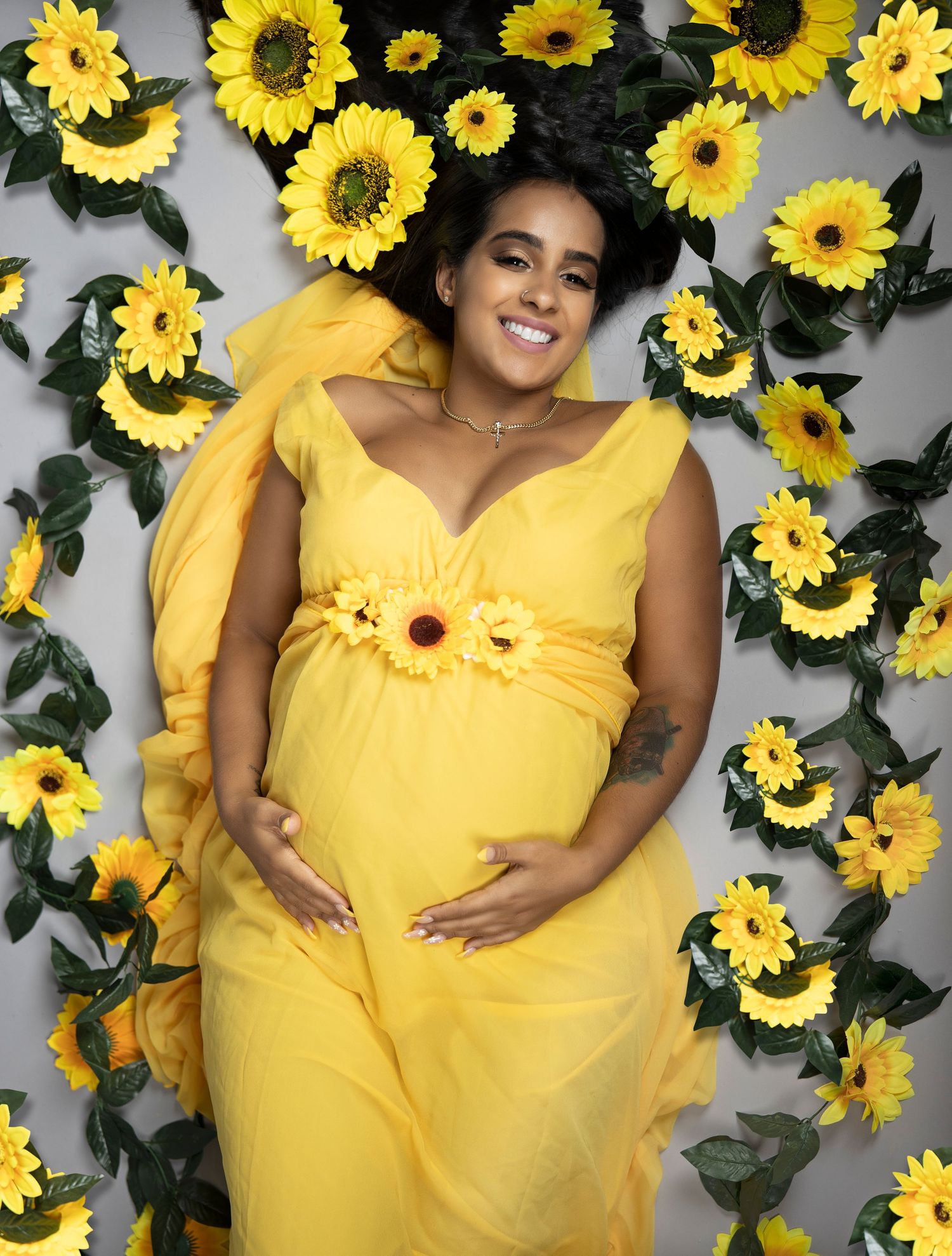 A radiant yellow maternity photo featuring a flowy dress surrounded by vibrant sunflowers against a gray backdrop.