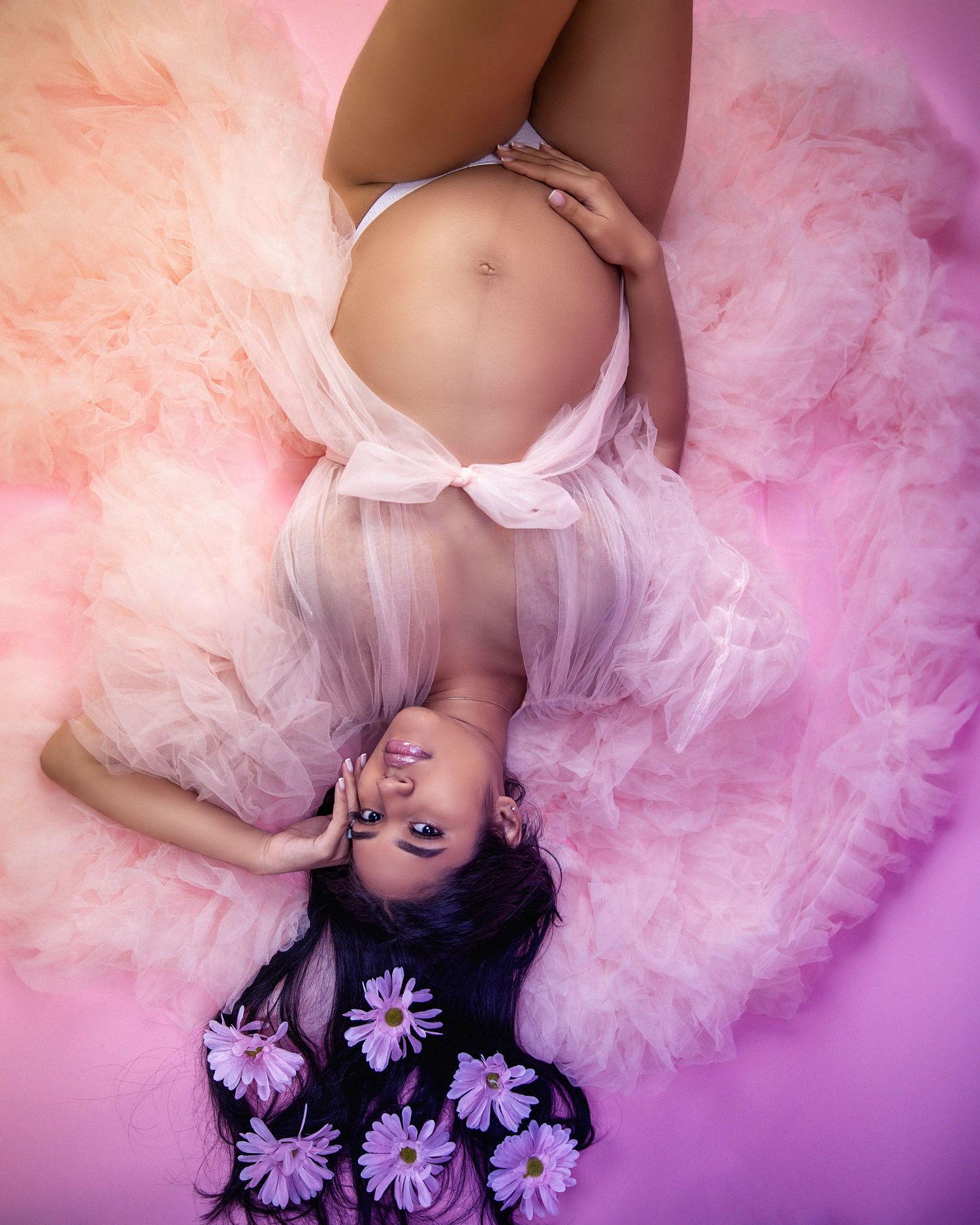 Artistic maternity photo with soft pink and purple tones featuring floral decorations and flowing tulle fabric on the ground.