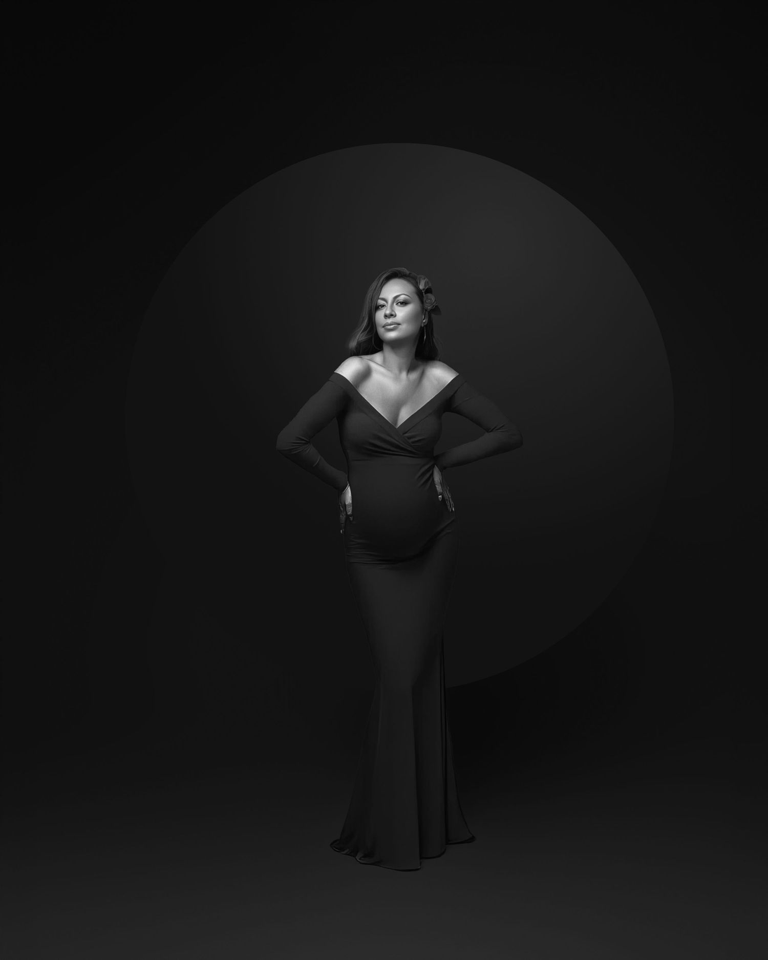 Dramatic black and white maternity portrait of a woman in a fitted gown posing against a dark circular backdrop.