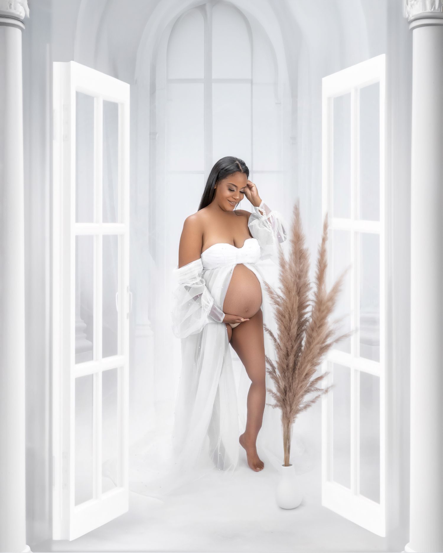 A maternity photo showing an elegant figure in white against a bright interior with pampas grass decor and open doors.