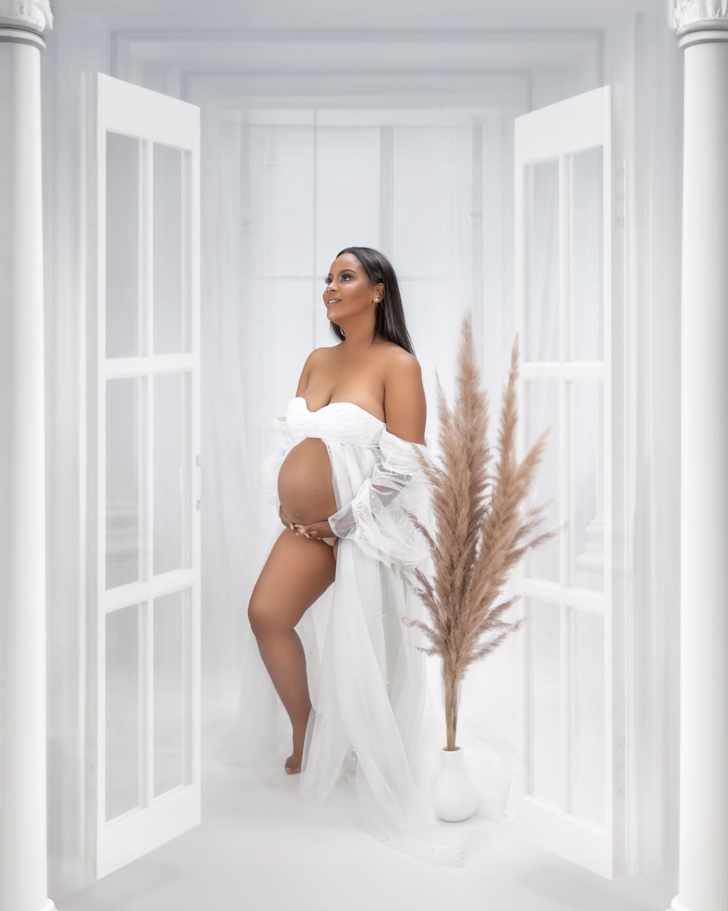 Elegant maternity photo featuring an expectant mother in a flowing white gown against a bright minimalist backdrop.