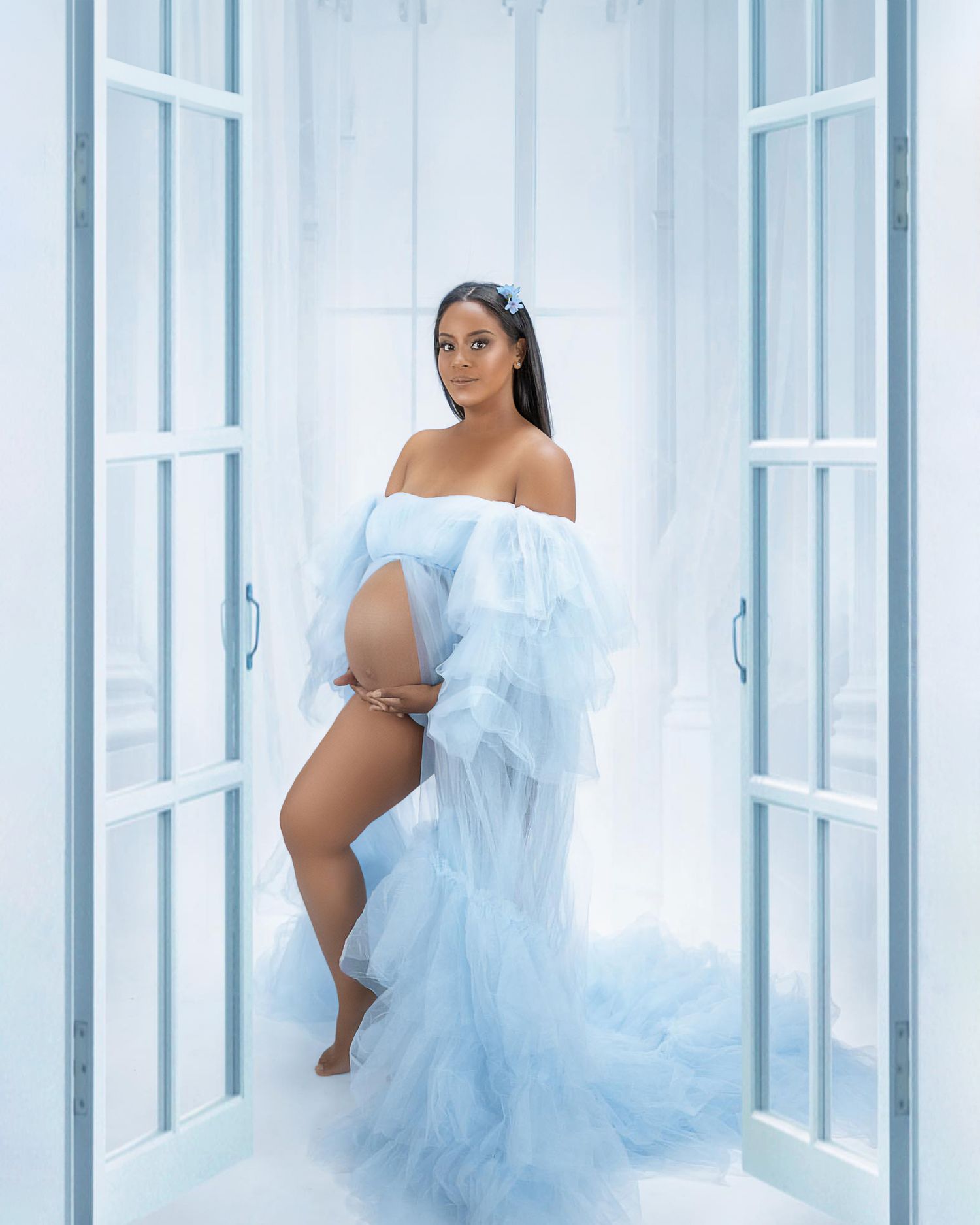 Ethereal maternity photo featuring a flowing light blue tulle gown against white French doors.