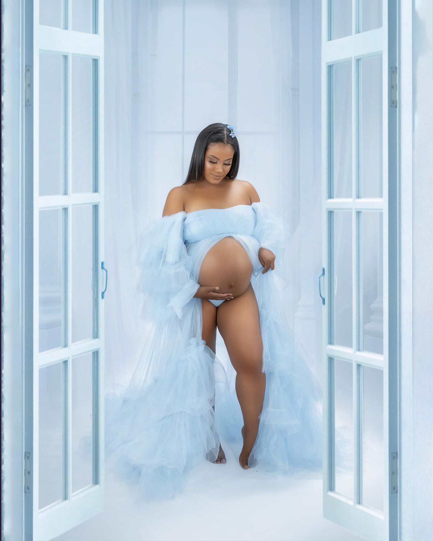 A maternity photo shoot in a white room with ethereal flowing fabric and soft lighting.