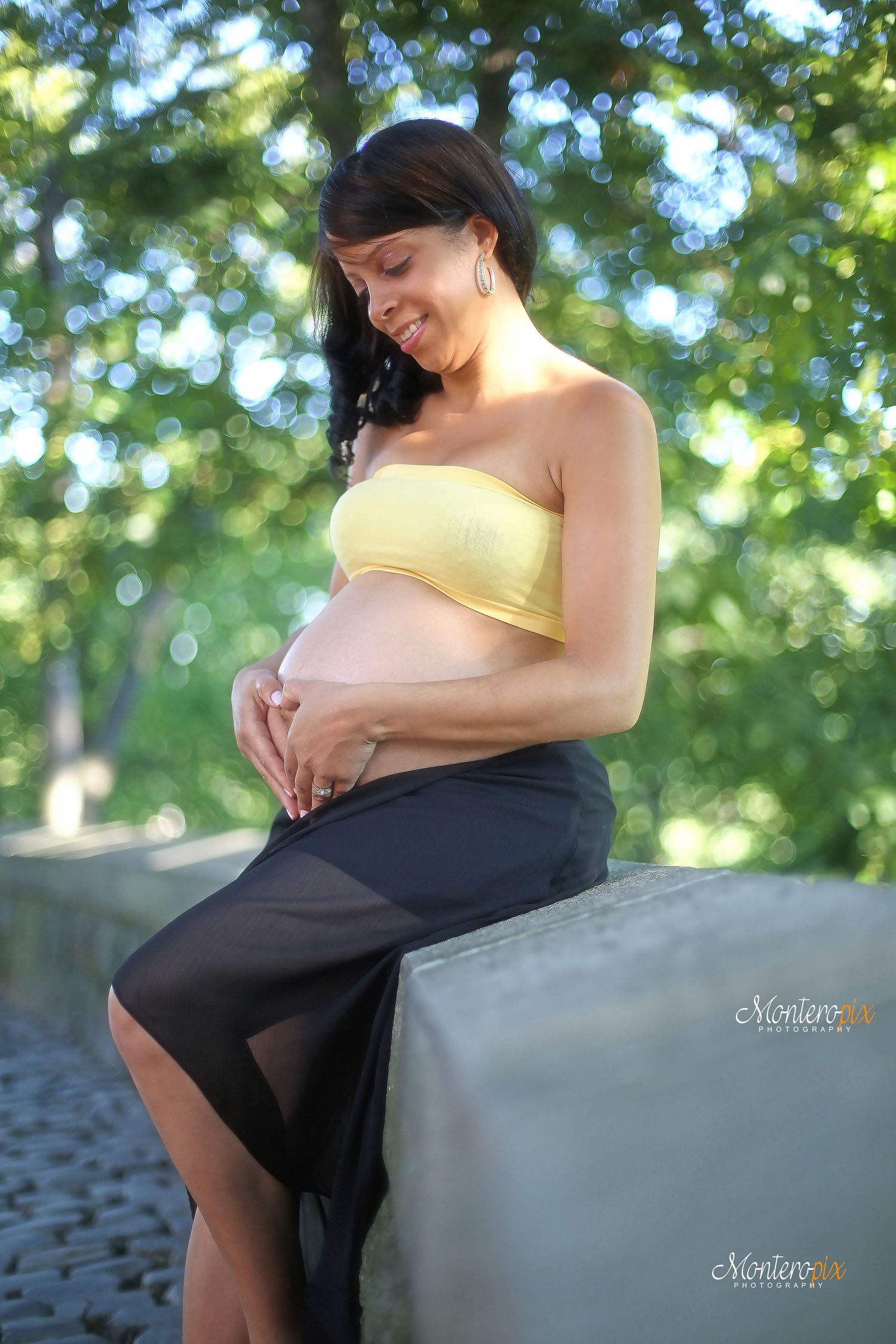 Expectant mother in yellow top sits peacefully on stone wall in dappled sunlight during outdoor maternity photoshoot.