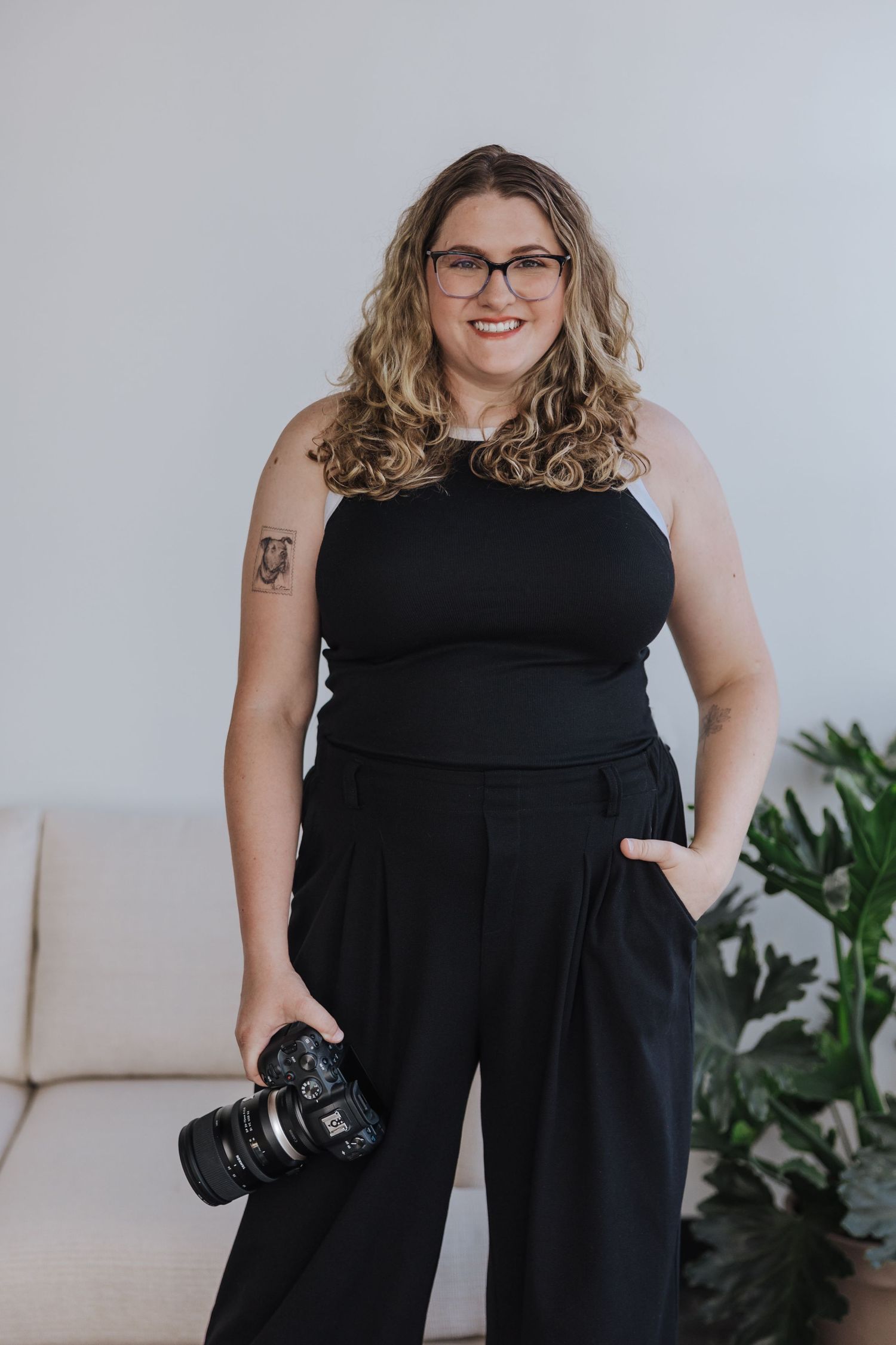Photographer wearing black jumpsuit and glasses holds professional camera while posing against light background.