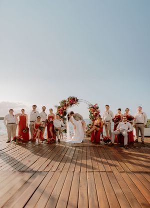 Wedding party in red and white attire poses on wooden deck by the ocean.