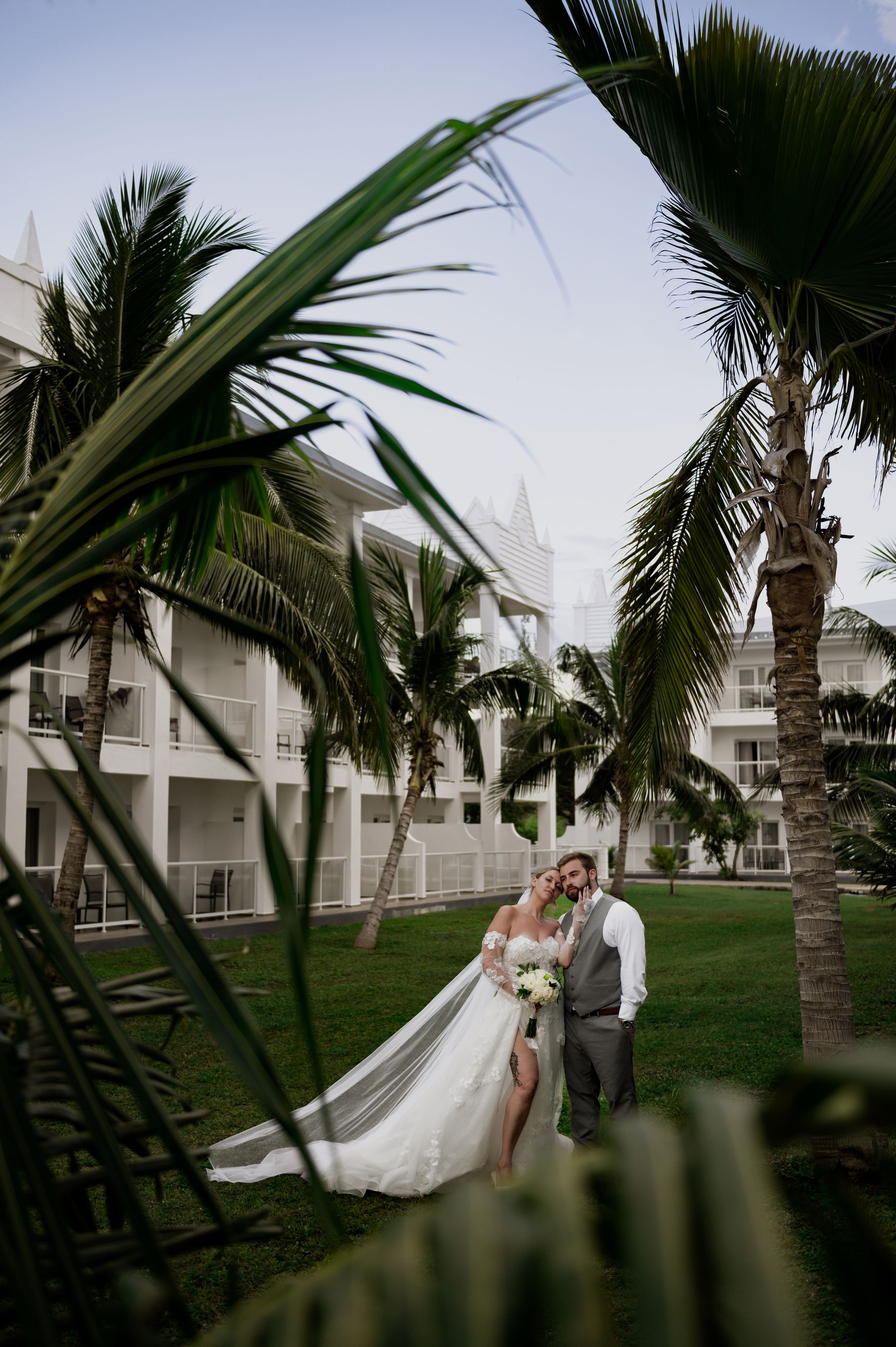 Bride and groom pose among tropical palm trees at a white resort venue on their wedding day.