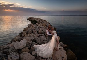Bride and groom embracing on rocky shoreline at sunset, with calm ocean waters and colorful sky in background.