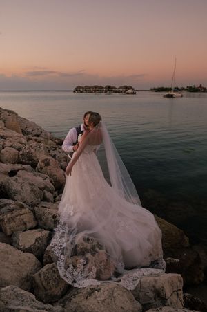 Bride and groom embrace on rocky shoreline at sunset, with lace wedding gown and veil flowing, ocean and overwater bungalows behind.