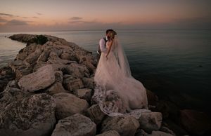 Bride in white gown and veil stands on rocky shoreline at sunset, gazing at calm ocean waters.