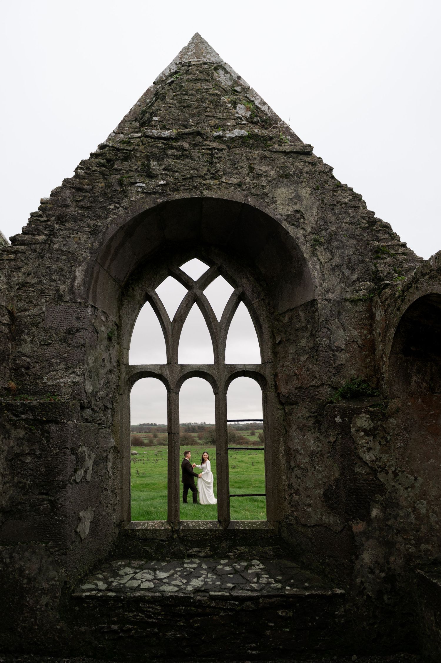 Ancient stone ruins with gothic arched windows frame a scenic countryside view.
