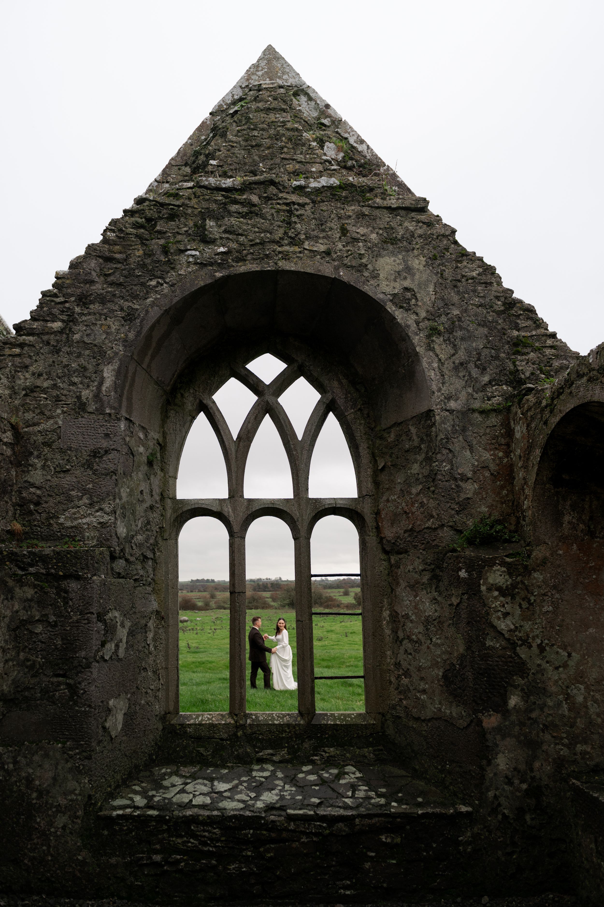 Ancient stone ruins with gothic arched windows frame a scenic countryside view.