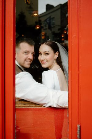 A wedding couple poses behind bright red window frames creating a striking contrast.