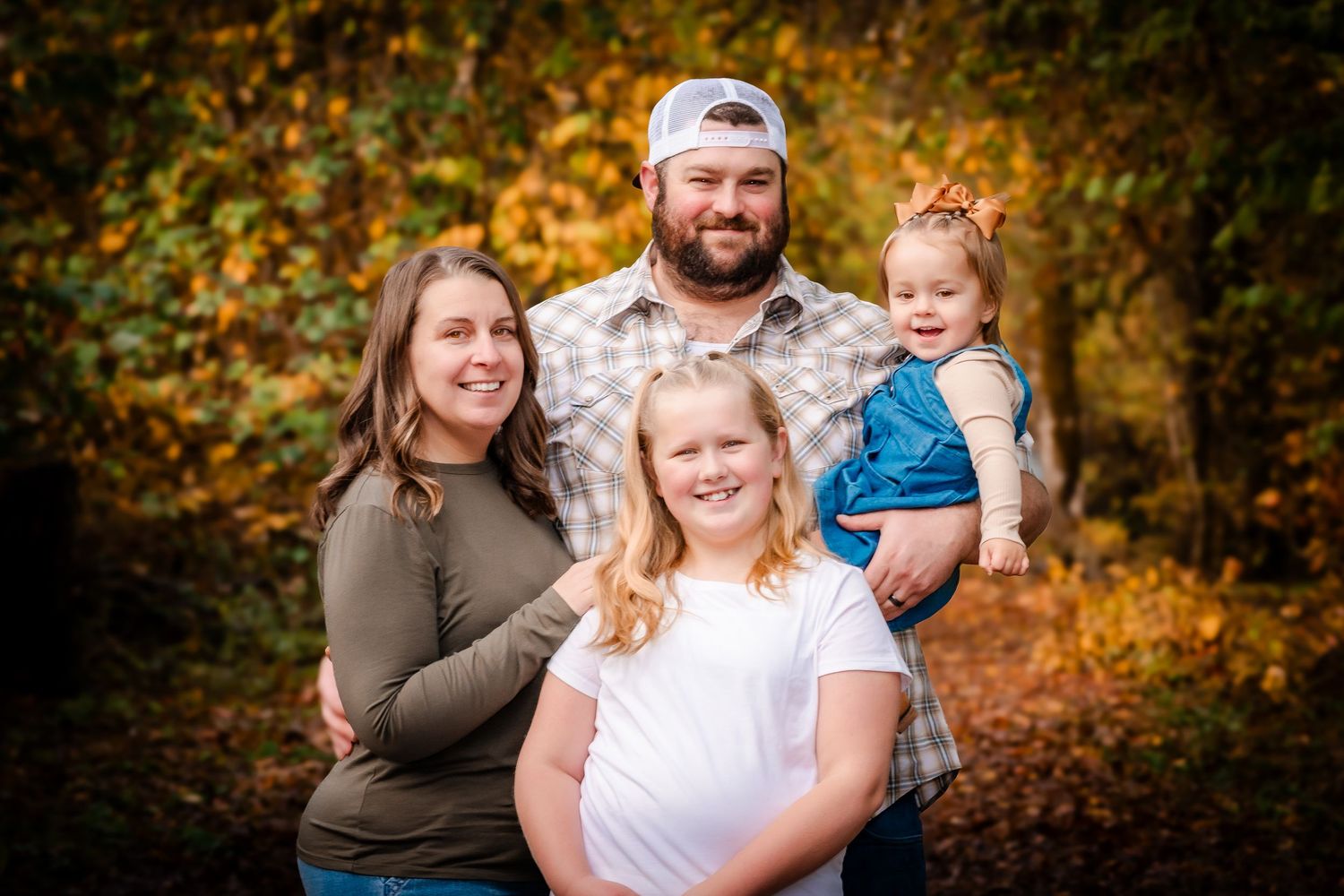 Family of four posing outdoors in autumn foliage, parents with two young daughters smiling in fall setting.