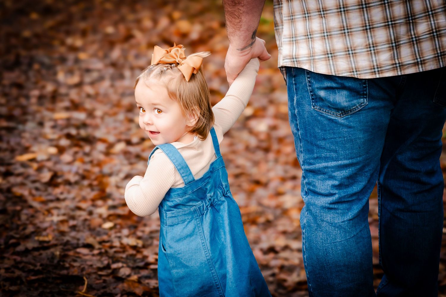 A toddler girl in blue overalls holds her father's hand on a leaf-covered autumn path.